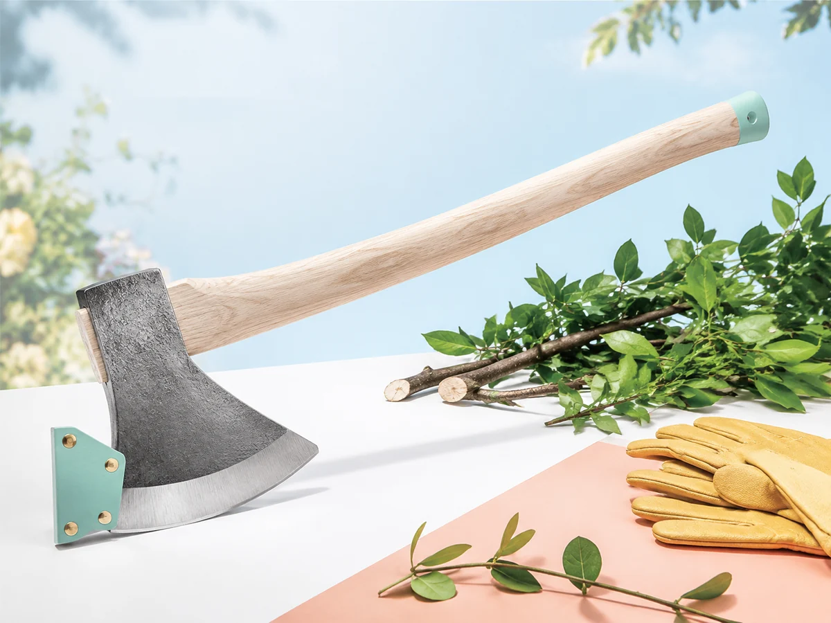 Brush-clearing garden axe with a broad forged head, edge guard, and ash handle beside fresh-cut twigs.