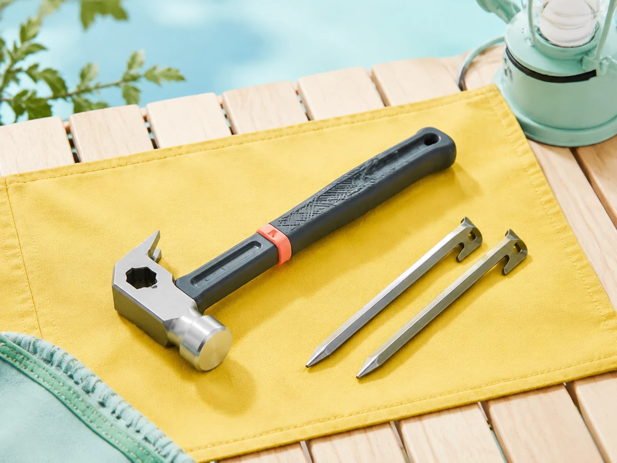 Compact camping hammer with a forged steel head, peg puller, coral grip, and tent stakes on a bright field mat.